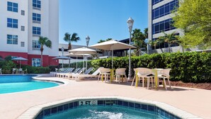 A swimming pool with lounge chairs and umbrellas, surrounded by a landscaped area with hedges and palm trees. - Hilton Garden Inn Tampa Airport Westshore (Tampa)