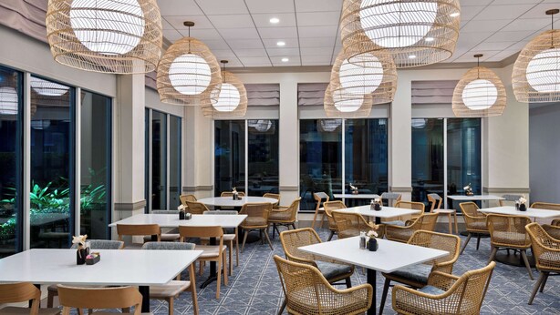 A restaurant with round tables and wicker chairs, large windows, and hanging wicker lamps. - Hilton Garden Inn Tampa Airport Westshore (Tampa)