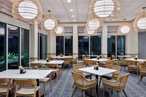 A restaurant with round tables and wicker chairs, large windows, and hanging wicker lamps. - Hilton Garden Inn Tampa Airport Westshore (Tampa)