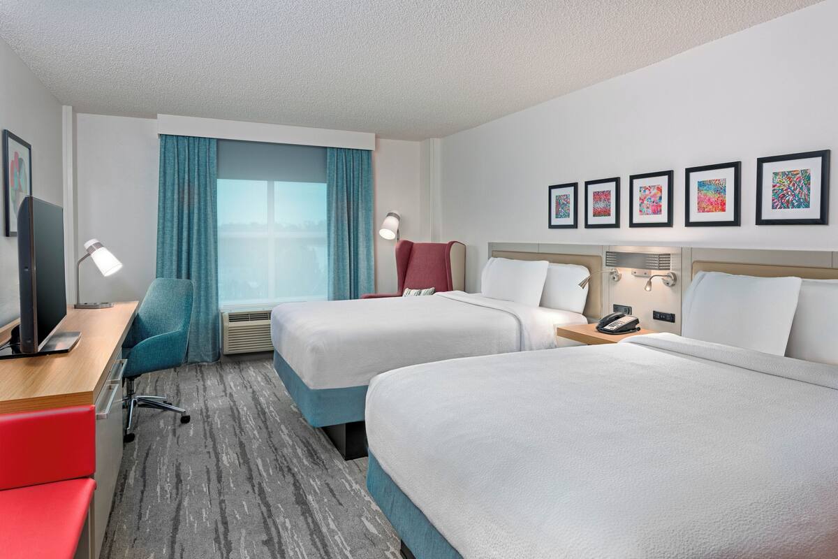 A hotel room with two beds, a desk with a computer, a TV, and a window with curtains.
