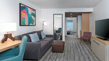 A modern hotel room with a grey sofa, teal armchair, and a large painting on the wall.