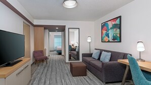 A modern hotel room with a flat-screen TV, a sofa, a chair, a small table, and a painting on the wall. - Hilton Garden Inn Tampa Airport Westshore (Tampa)