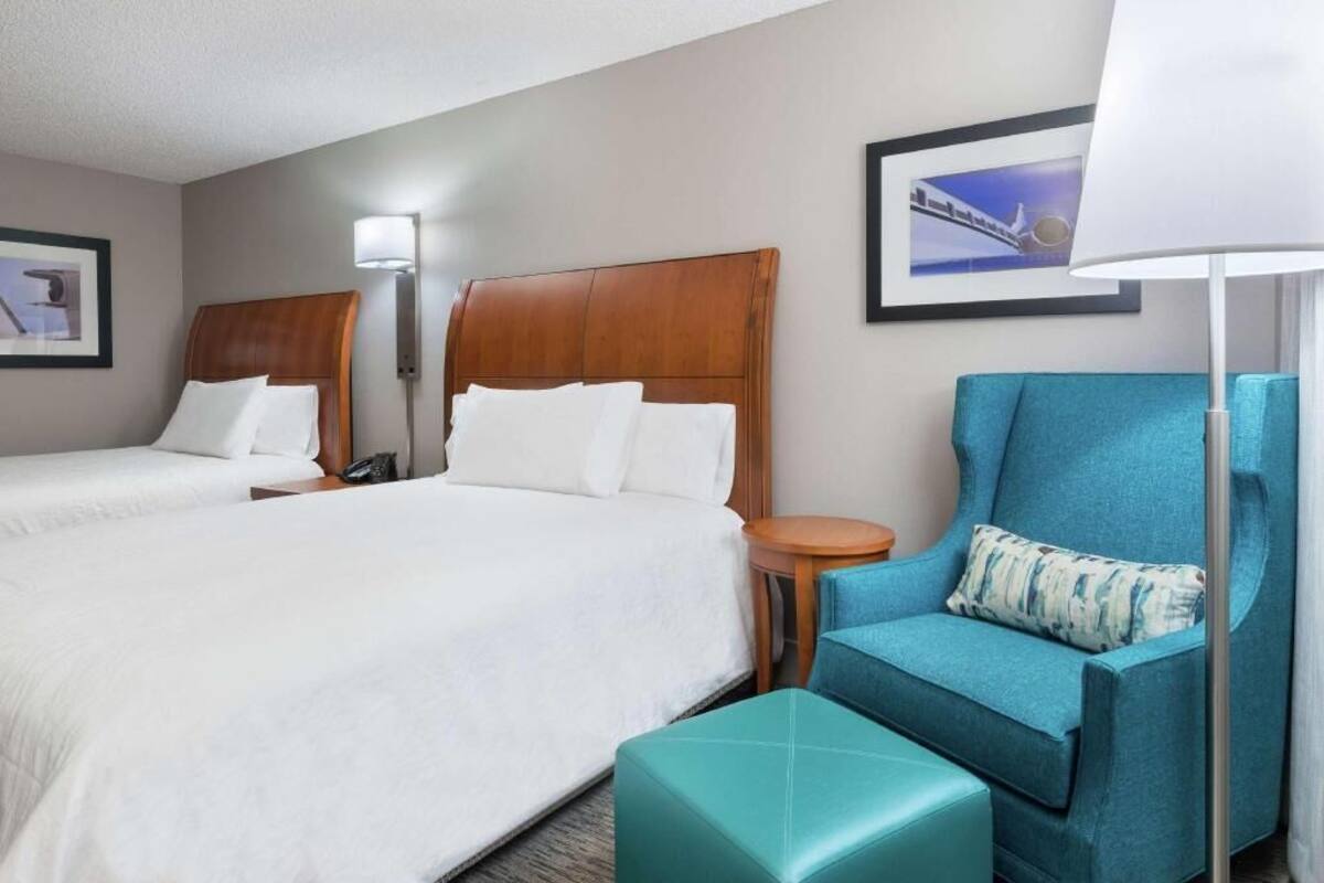A hotel room with two beds, a blue armchair, a small round table, and a lamp.