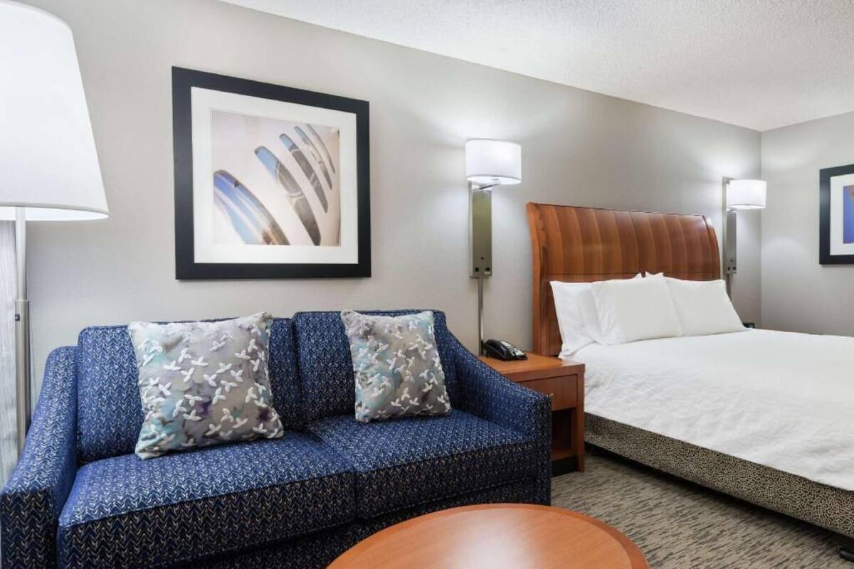 A hotel room with a bed, a sofa with patterned cushions, a small round table, and two wall-mounted lamps.
