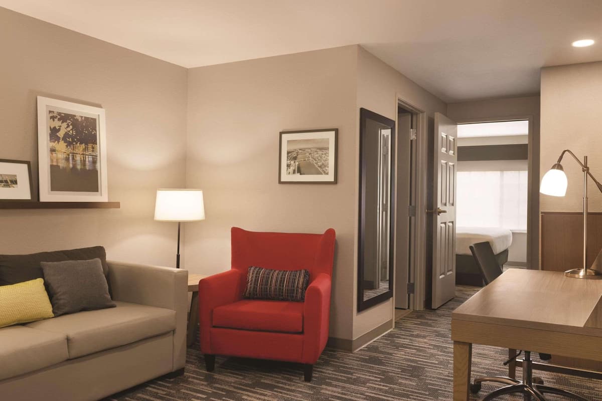 A hotel room with a sofa, a red armchair, a desk, and a lamp. There are framed pictures on the wall and a door leading to another room.