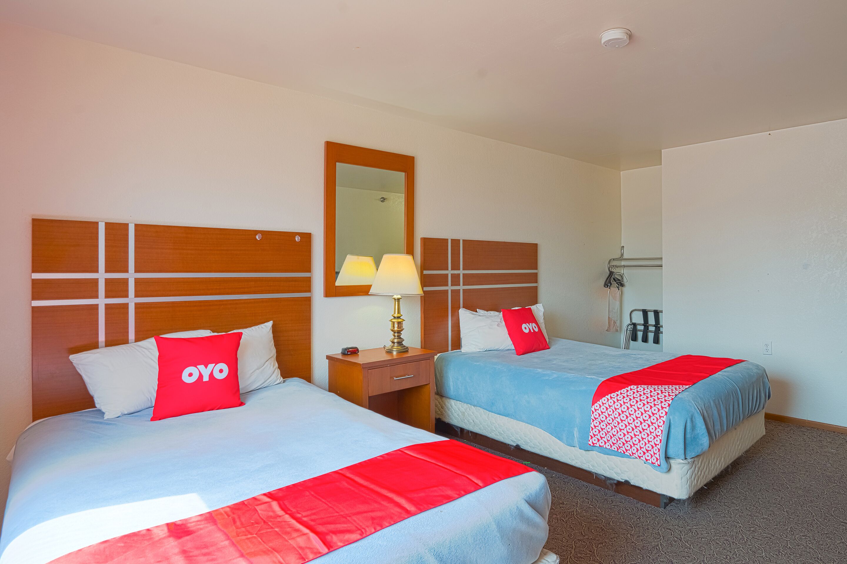 A hotel room with two beds, each with a red and white bedspread, a wooden headboard, a nightstand with a lamp, and a mirror on the wall.