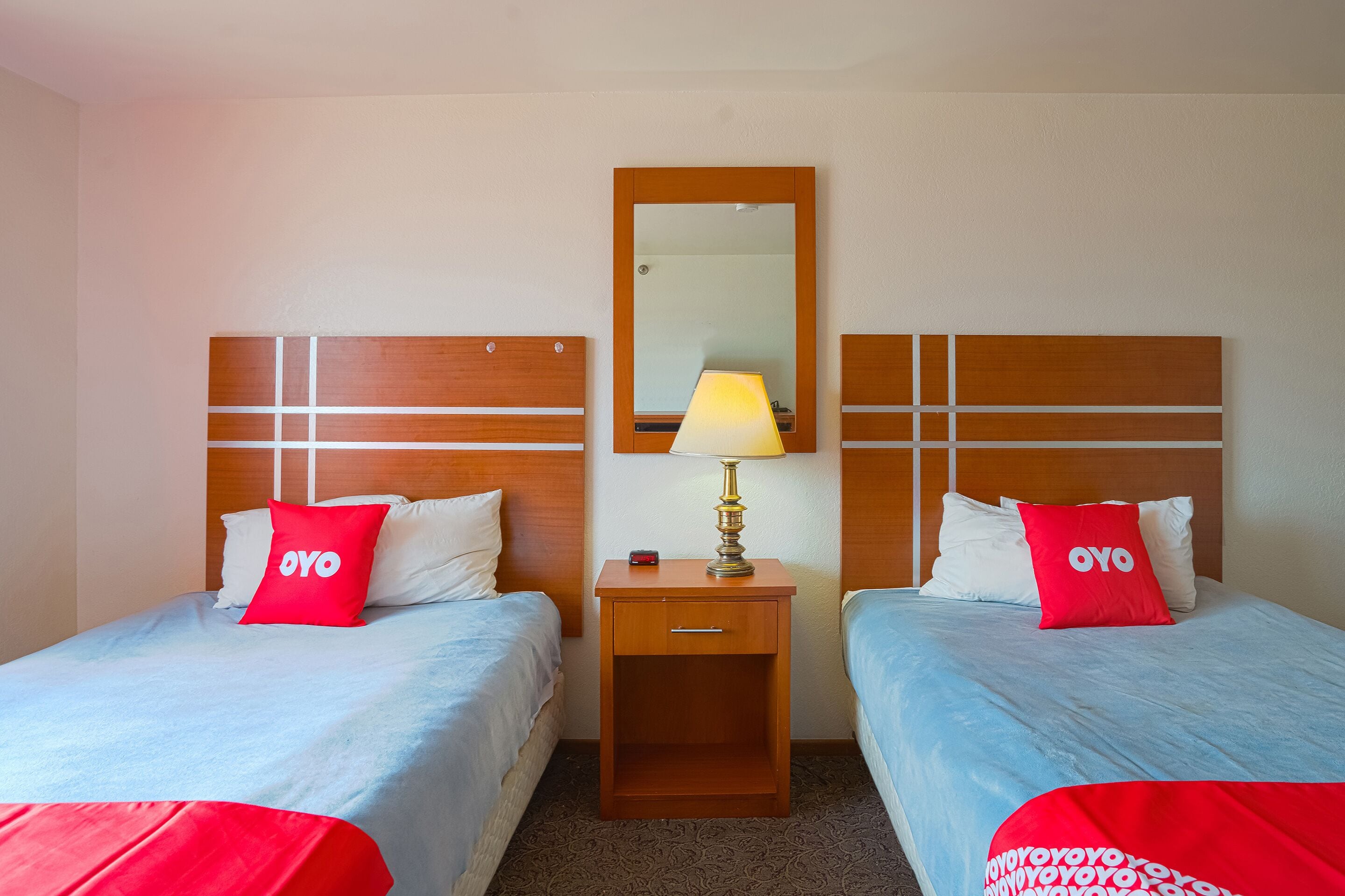 A hotel room with two beds, each with a red pillow and a blue bedspread. There is a wooden nightstand with a lamp between the beds.