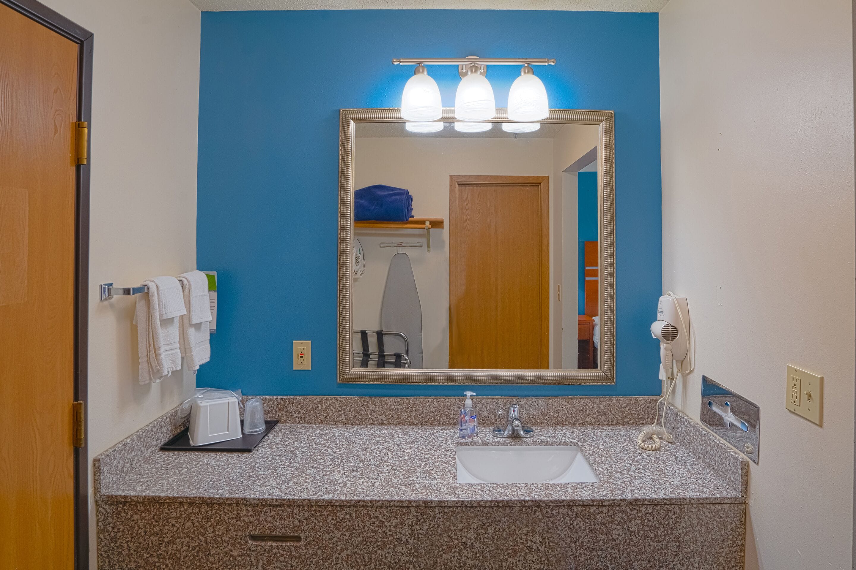 Une salle de bain avec un mur d’accent bleu, un grand miroir, un lavabo avec un robinet, un sèche-cheveux et un porte-serviettes.
