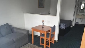 A compact room with a grey sofa, a small wooden table with a chair, a single bed, and a mirror above the bed. - Relax Inn Dover Near Speedway (Dover)