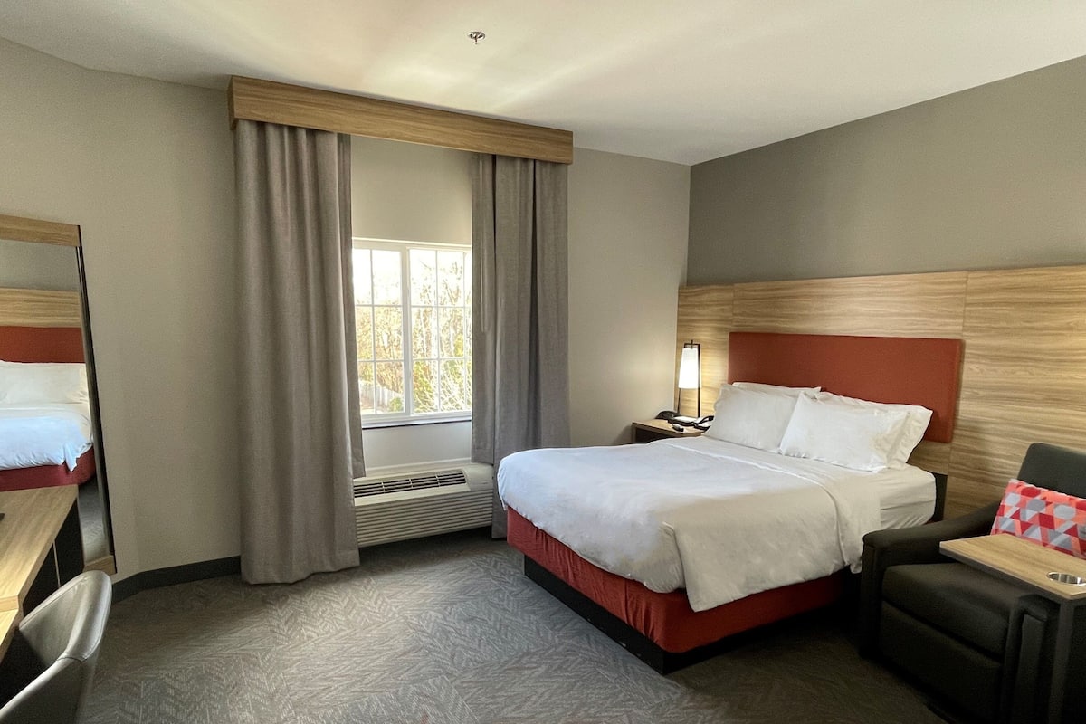 A hotel room with a bed, a desk, a chair, a nightstand with a lamp, and a window with curtains.