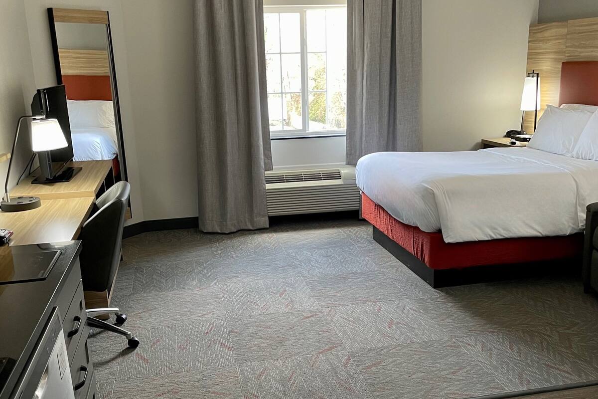 A hotel room with a bed, a desk with a lamp, a dresser, a mirror, and a window with curtains.