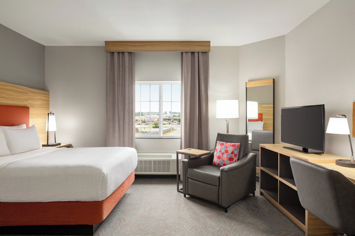 Une chambre d’hôtel avec un lit, un bureau avec une télévision, une chaise et une fenêtre avec des rideaux.