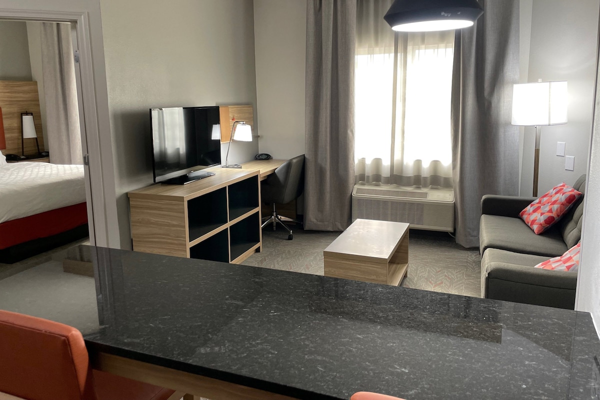 Quarto de hotel com cama, escrivaninha com computador, sofá, mesa de centro e uma janela grande com cortinas.