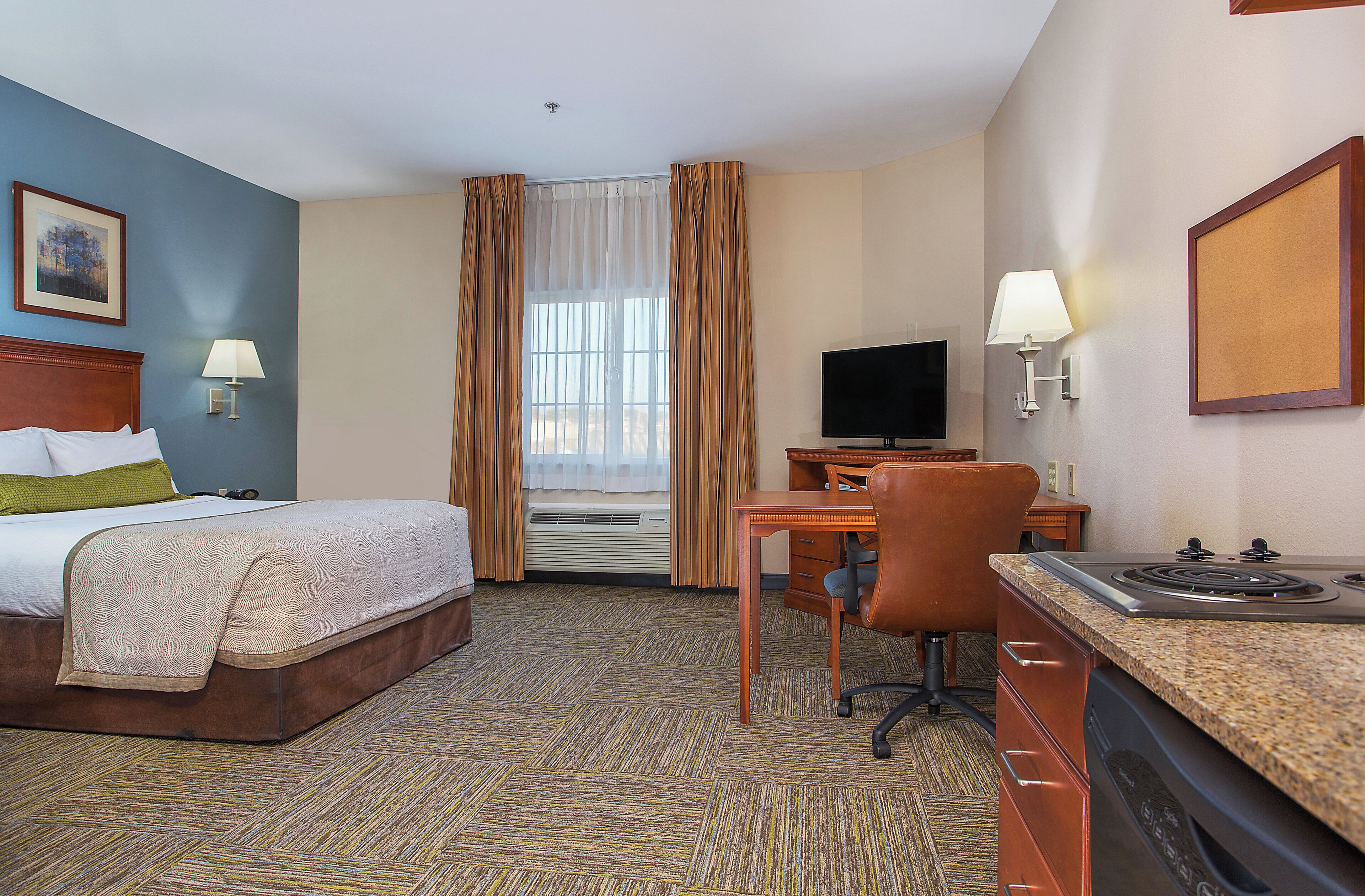 A hotel room with a bed, desk, chair, television, and kitchenette.