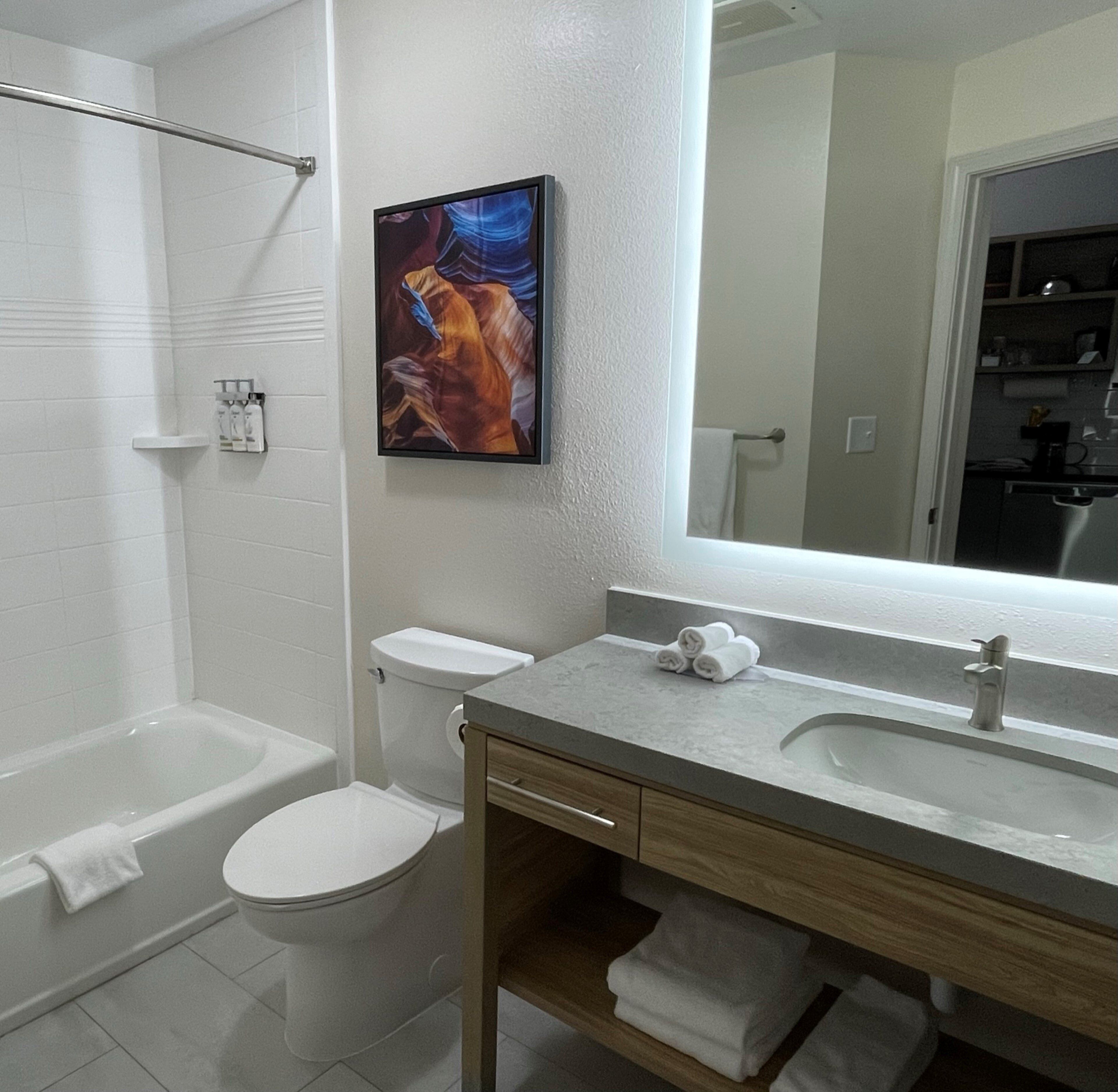 A modern bathroom with a bathtub, toilet, and sink. There is a painting above the sink and a large mirror above the sink.