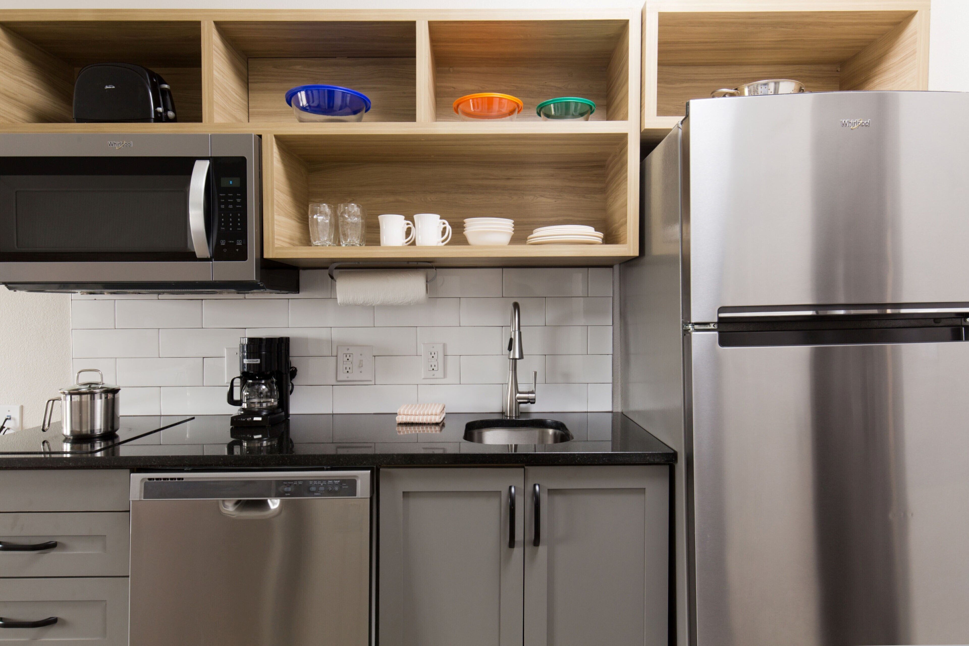 A modern kitchen with a built-in microwave, coffee maker, and dishwasher. There are wooden shelves above the counter holding bowls and cups.