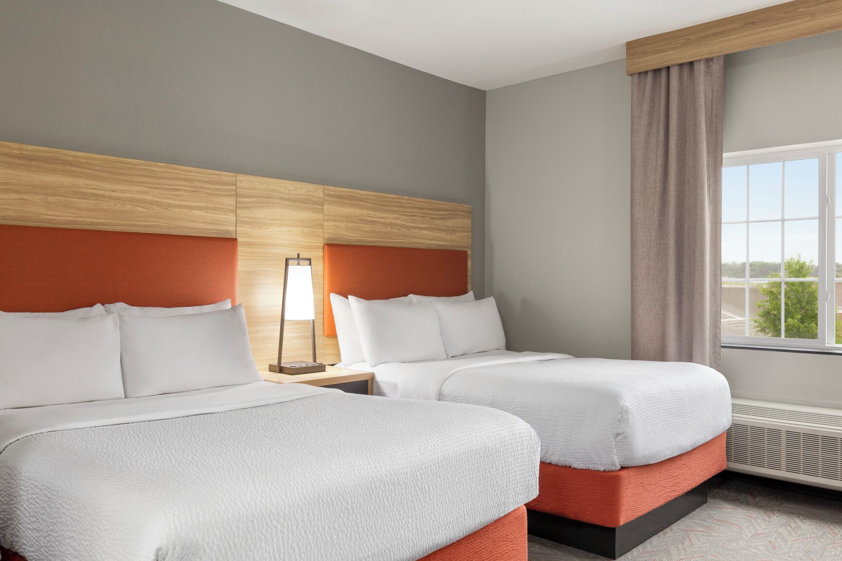 A hotel room with two beds, a nightstand with a lamp, a window with curtains, and a view of the outdoors.