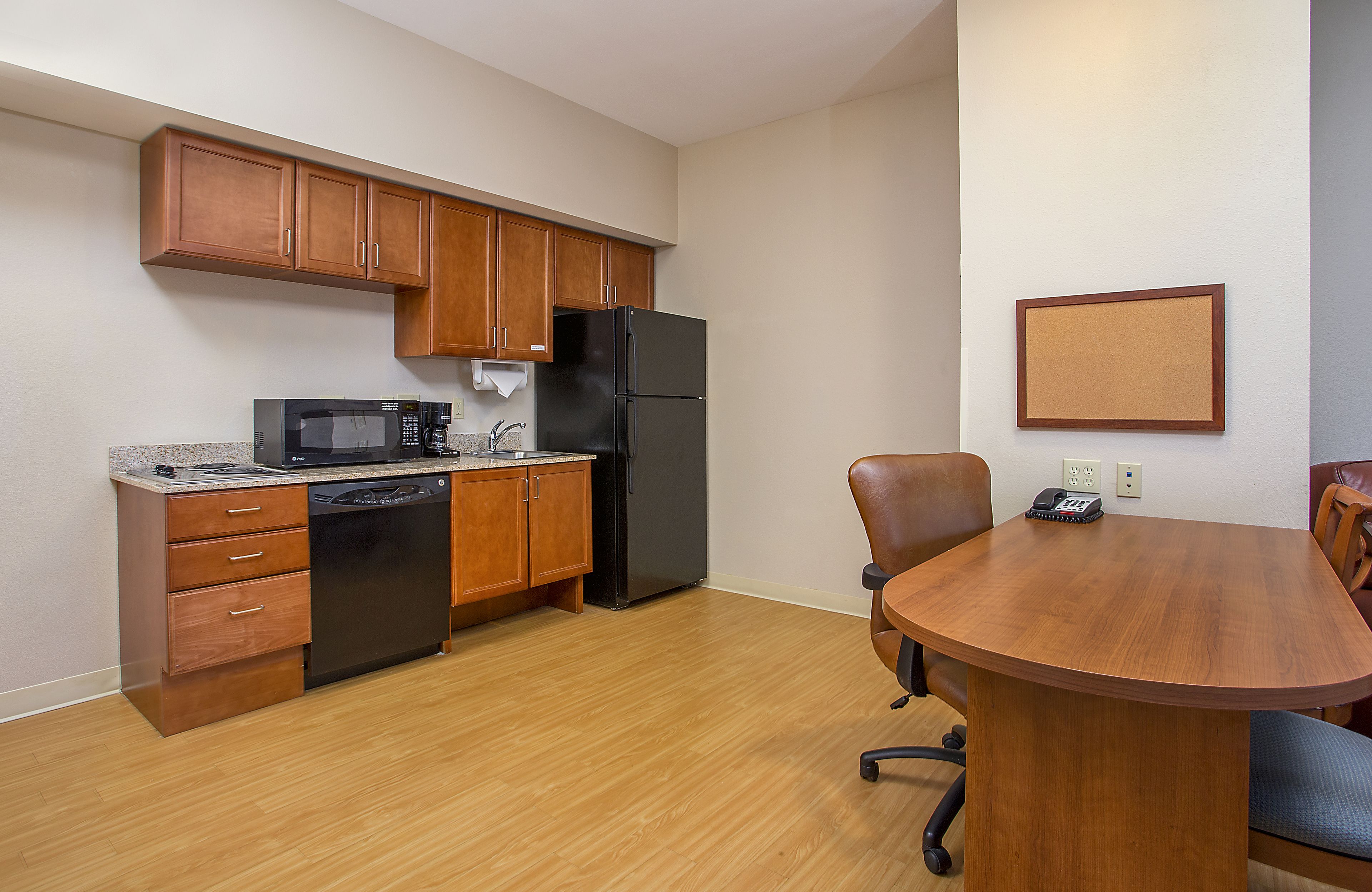 A compact kitchen with wooden cabinets, a black refrigerator, a microwave, and a dishwasher. There's a dining table with chairs and a corkboard on the wall.