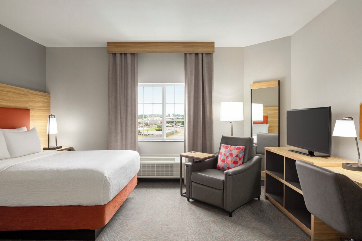 Une chambre d’hôtel avec un lit, un bureau avec une télévision, une chaise et une grande fenêtre avec des rideaux.