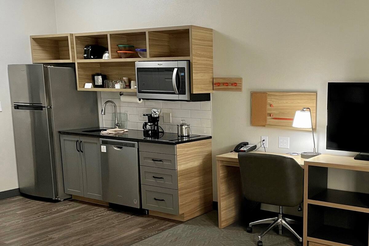A compact kitchen with a refrigerator, microwave, and coffee maker, a wooden desk with a chair, and a TV on a stand.