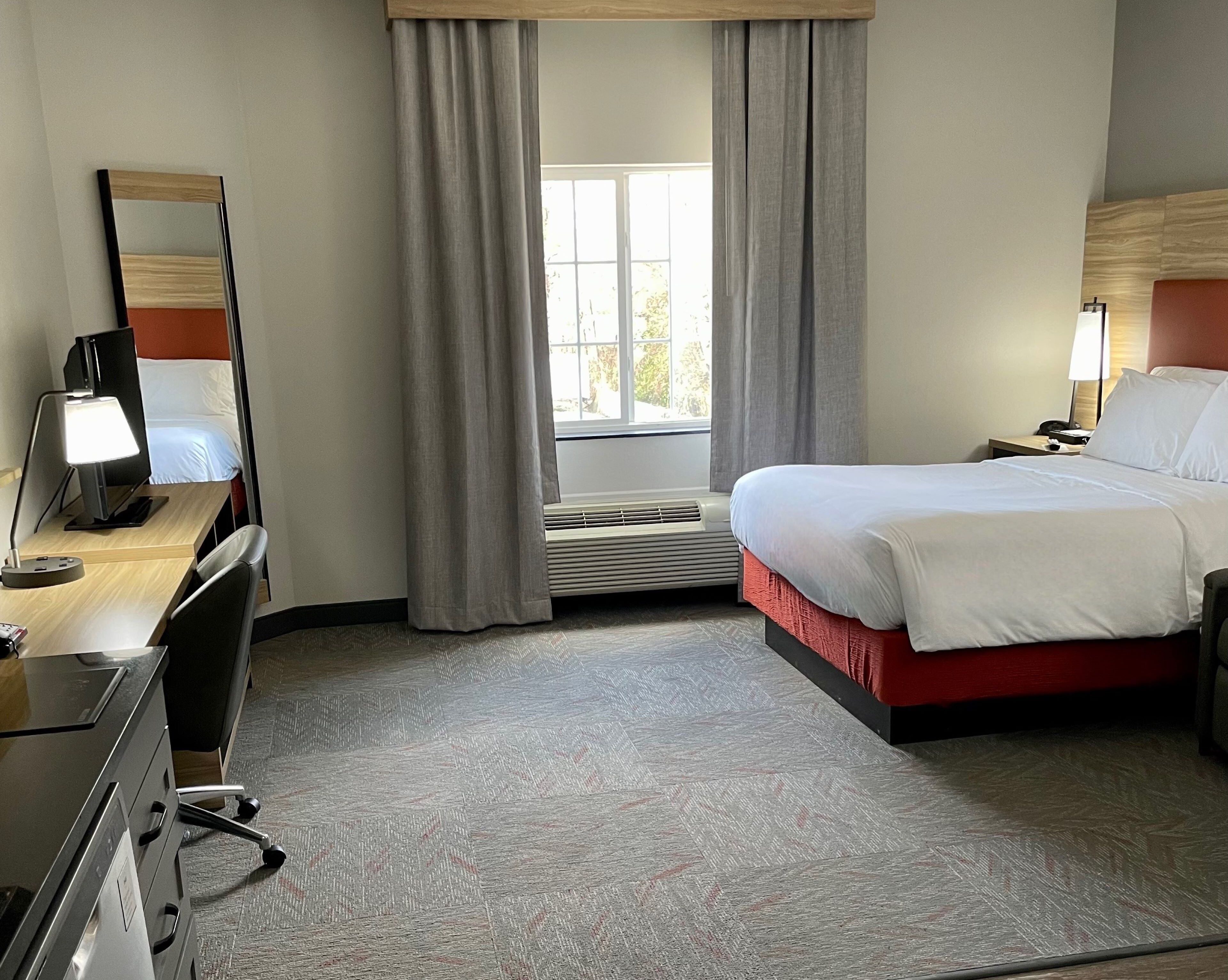A hotel room with a bed, a desk with a lamp, a dresser, a mirror, and a window with curtains.