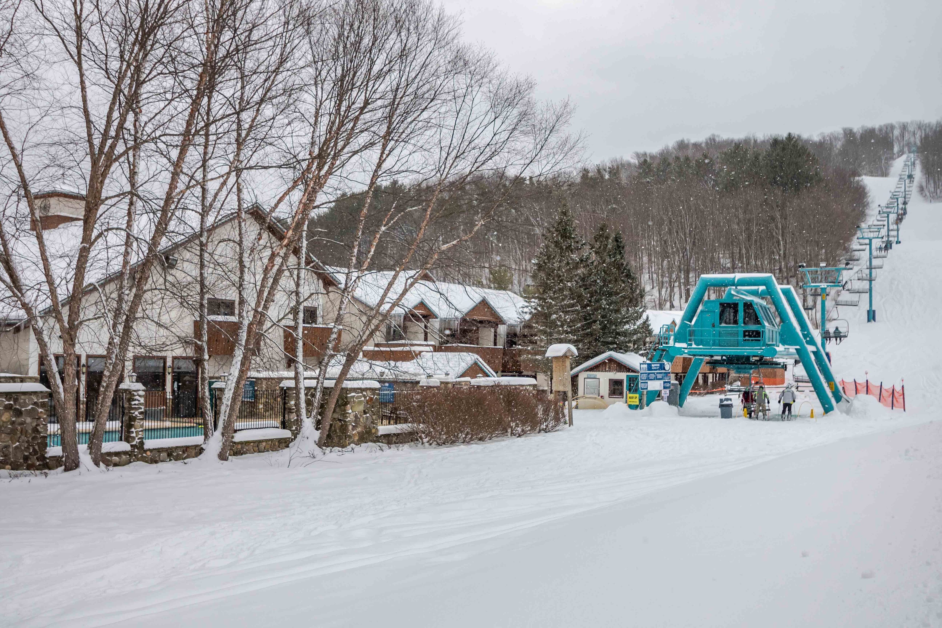 A snowy ski resort with a lift station and a residential area with houses and bare trees.