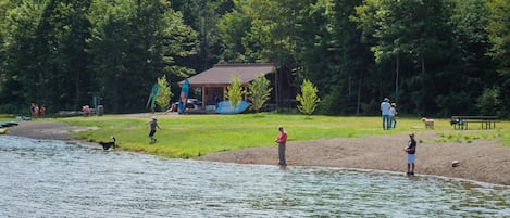Une scène au bord d’un lac, avec un chalet en bois, des zones herbeuses et des personnes près de l’eau.