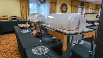 Un buffet composé de plats présentés dans des récipients couverts, d’un plateau contenant des couverts et d’une guirlande décorative accrochée au mur.