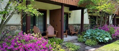 Um deck de madeira com duas cadeiras Adirondack, um muro de pedra e um jardim com flores roxas.