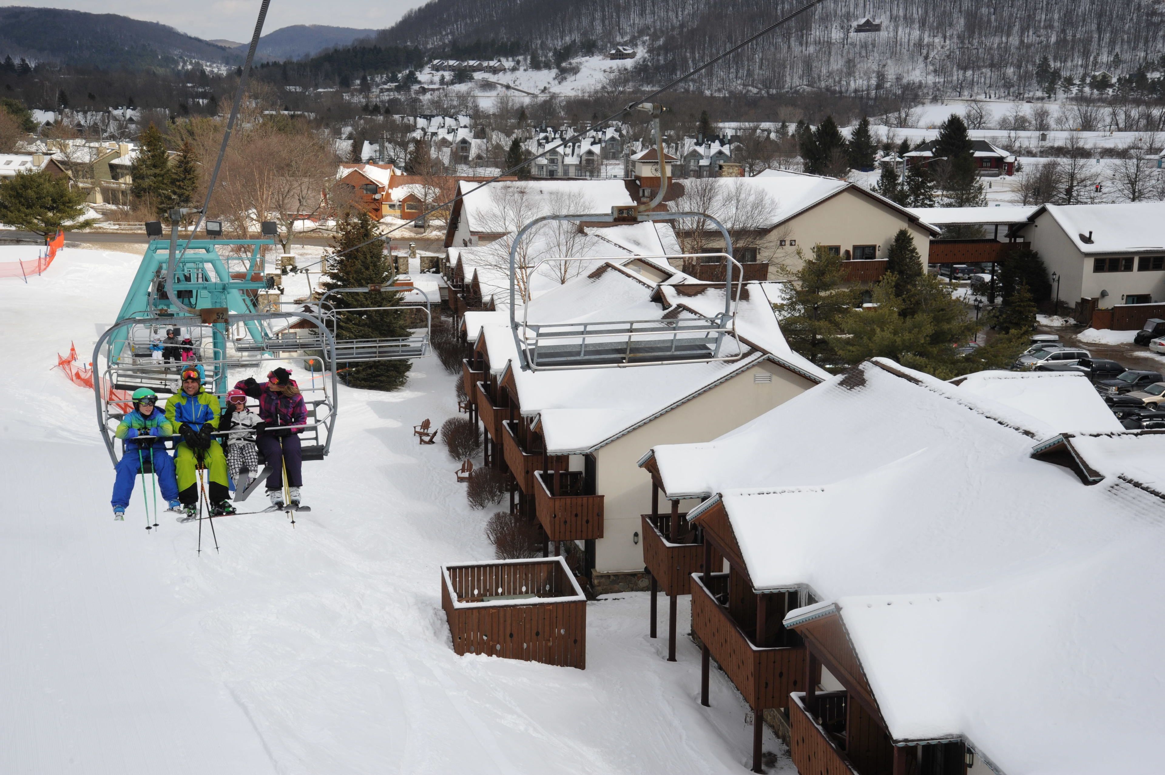 A ski lift with passengers, wooden buildings with snow-covered roofs, and a snowy landscape with houses and mountains in the background.