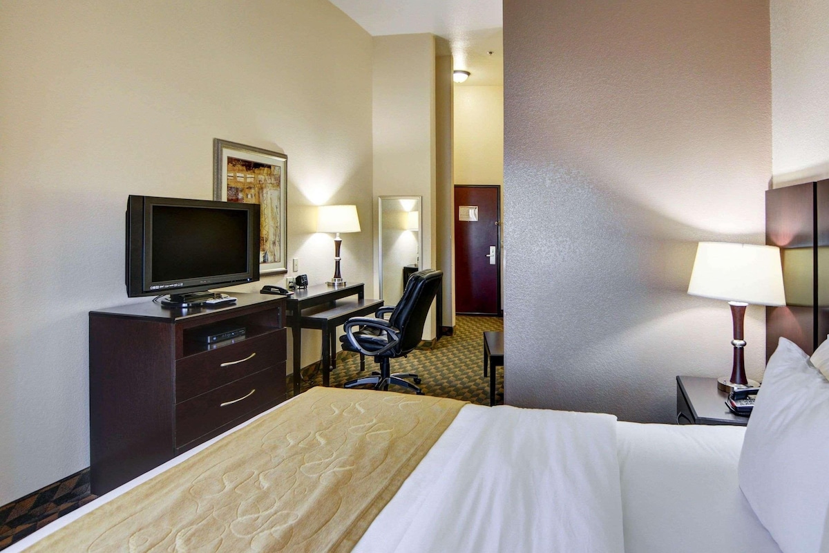 Habitación de hotel con una cama, un escritorio con televisor, dos sillas, una lámpara de escritorio y una puerta que da a otra habitación.