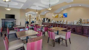 Un'area ristorazione di un hotel con diversi tavoli e sedie, un bancone della cucina con esposizioni di cibi e un televisore a parete.