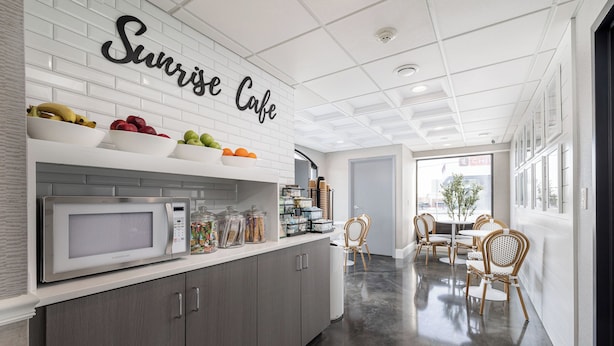 A modern kitchen with a white backsplash, a microwave, and a fruit bowl. There is a dining area with wicker chairs and a table. - Oakfield Hotel & Suites, Ascend Collection, Rochester-Pittsford NY (Rochester)