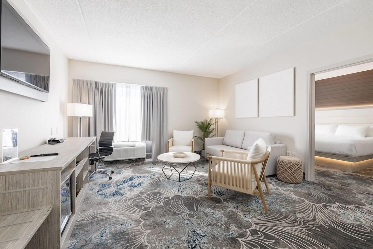 A hotel room with a desk, two chairs, a sofa, a bed, and a large window with curtains.
