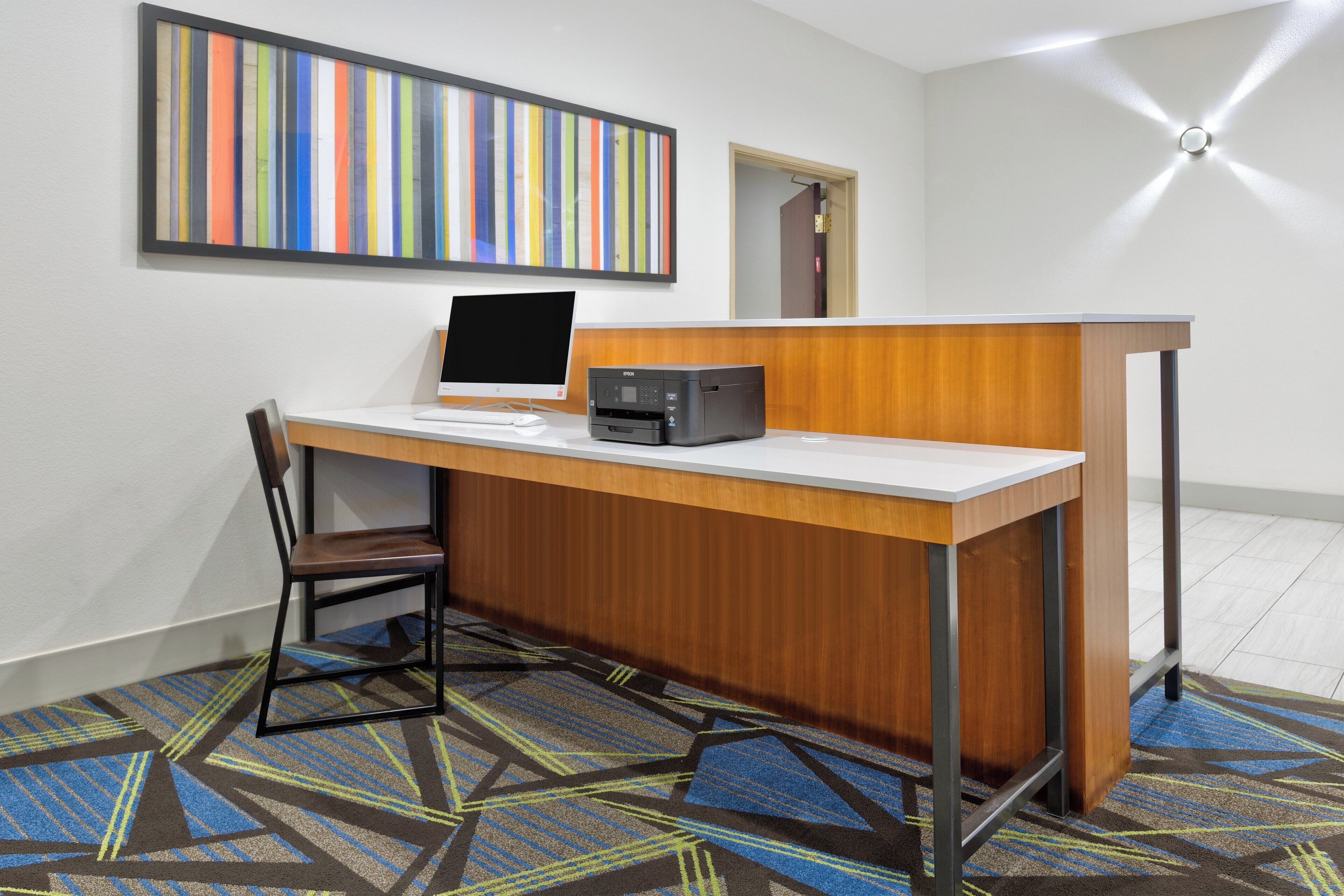 Taulell de recepció amb un ordinador, una impressora i una cadira en una sala amb un mosaic de moqueta i una obra d'art mural amb franges de colors.