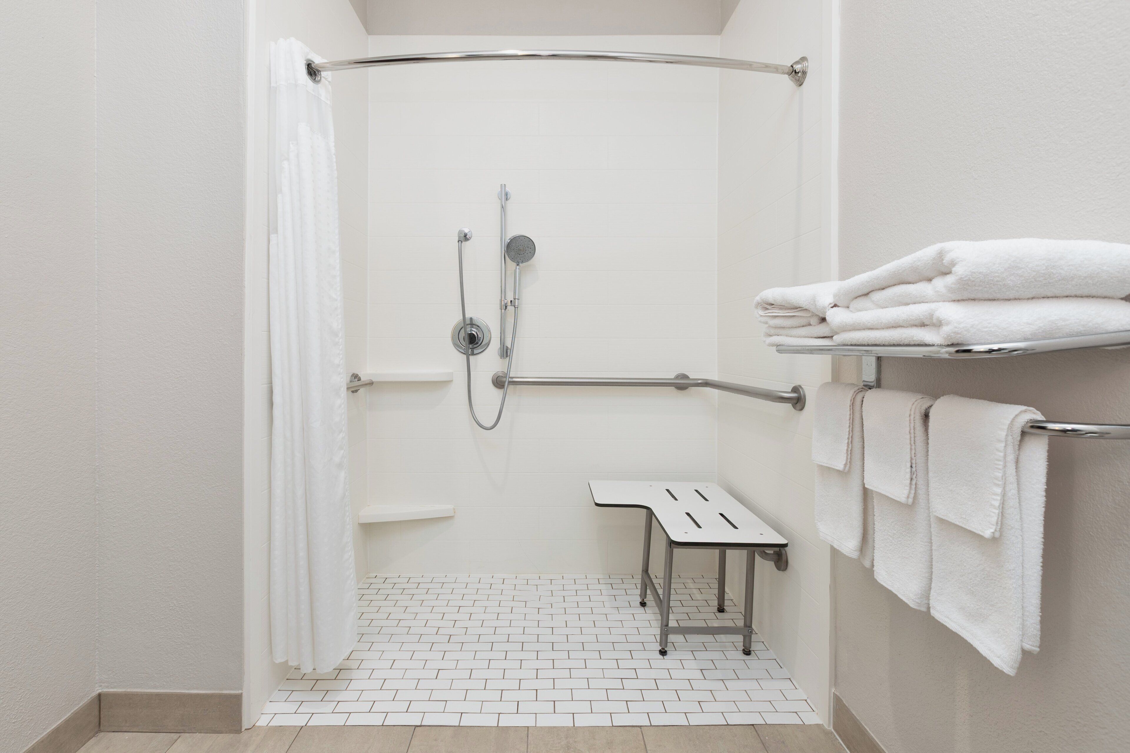 Un espace douche équipé d’une barre d’appui, d’un sol carrelé de couleur blanche et d’une étagère où sont pliés les serviettes.