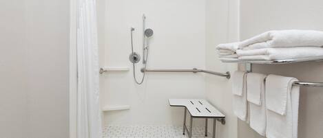 Un espace douche équipé d’une barre d’appui, d’un sol carrelé de couleur blanche et d’une étagère où sont pliés les serviettes.