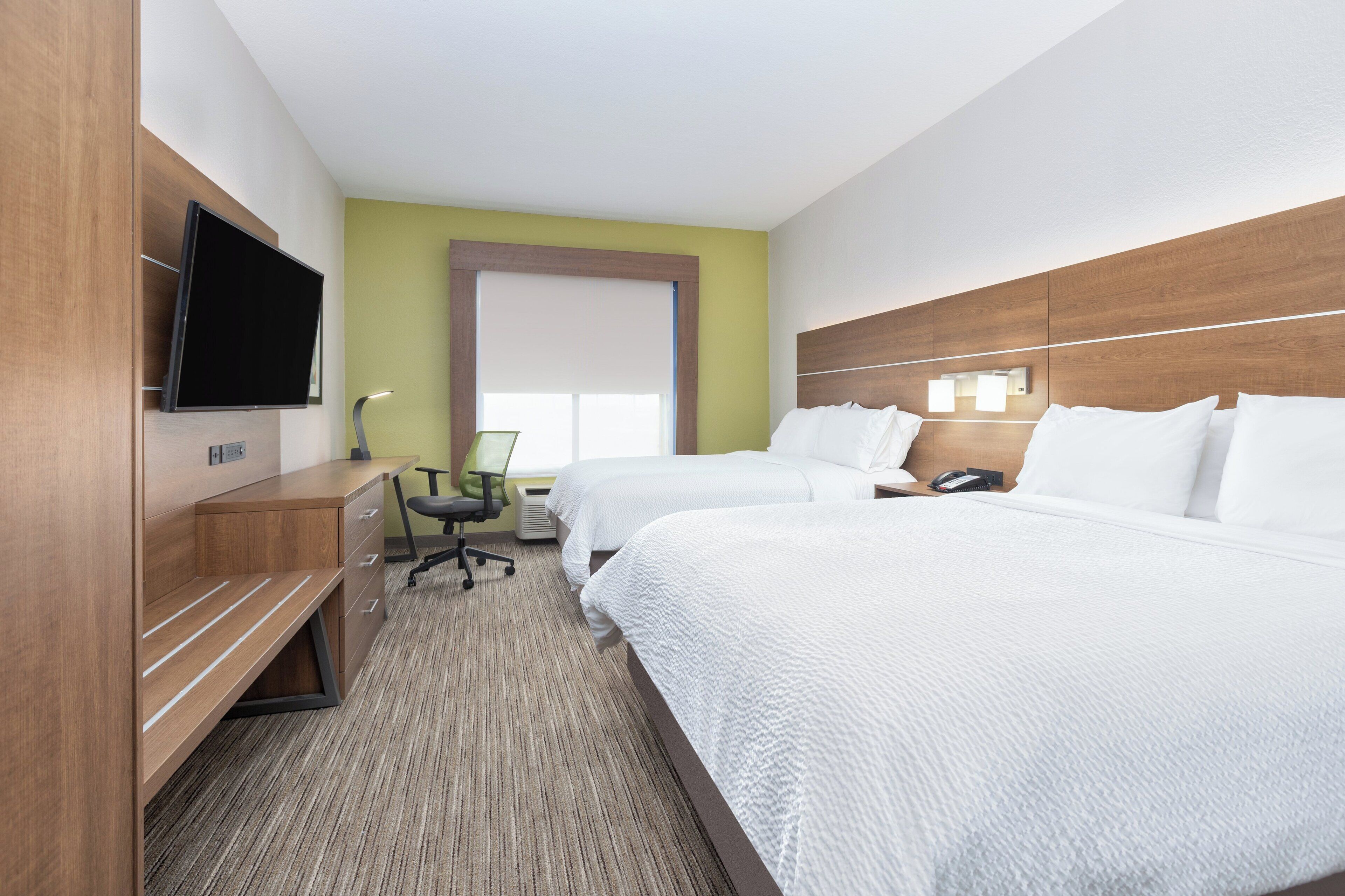 A hotel room with two beds, a desk, a chair, a flat-screen TV, and a window with blinds.