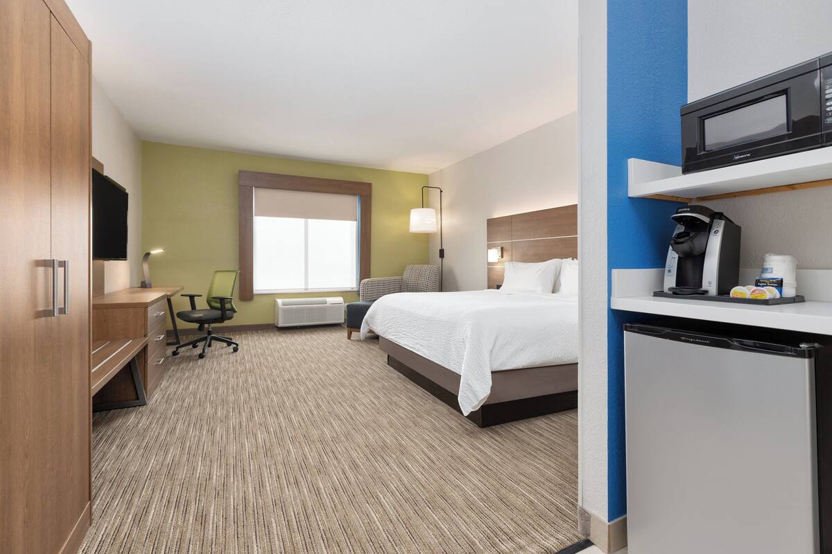 A hotel room with a bed, desk, chair, and a microwave on a shelf.