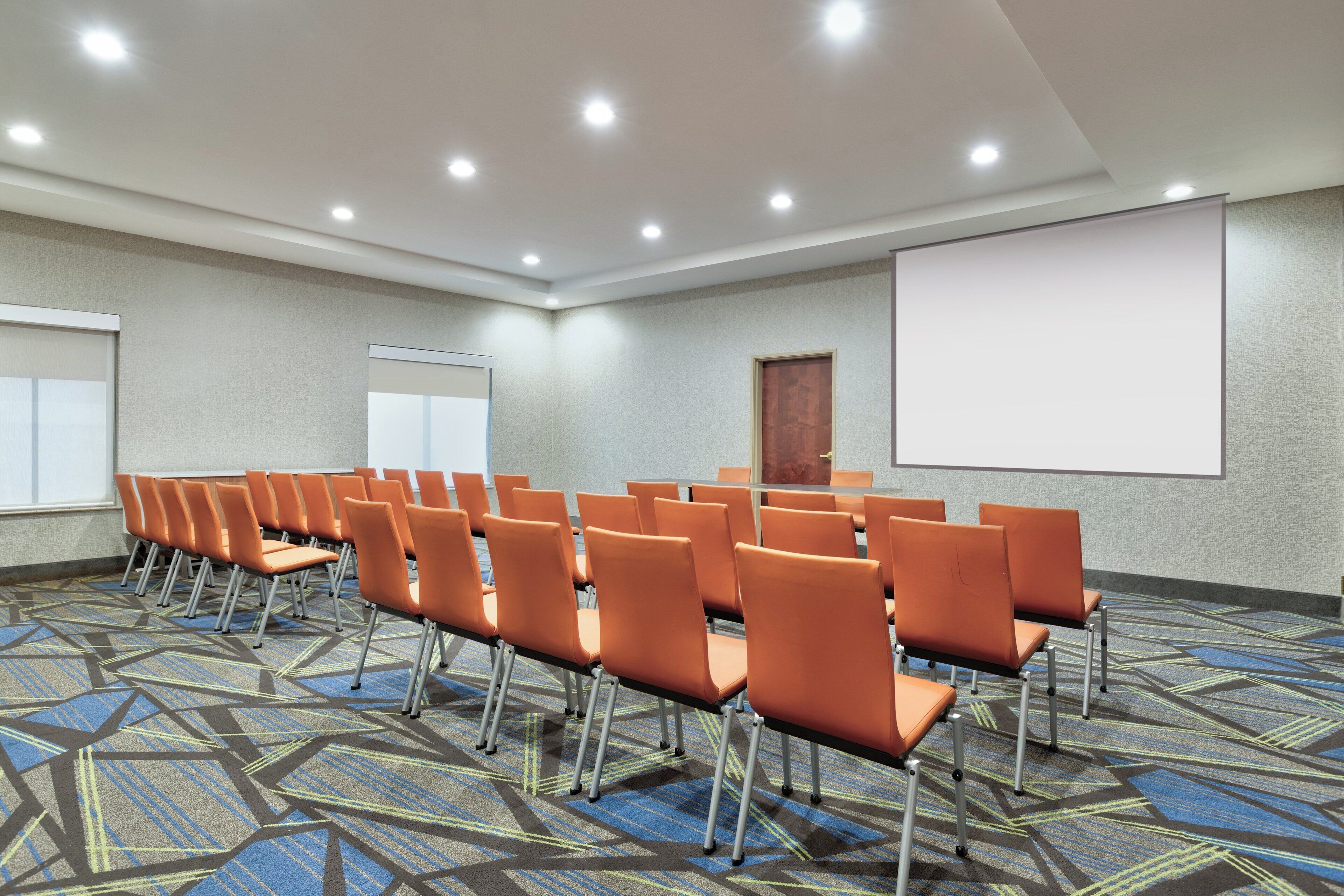 Une salle de conférence avec des rangées de chaises orange, un grand écran et une moquette à motifs.