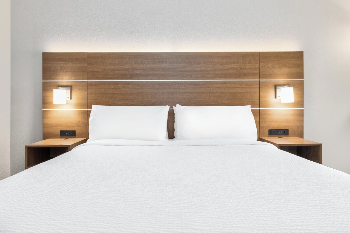 Un lit bien fait, avec une tête de lit en bois, deux tables de chevet dotées de lampes fixées au mur, et un mur blanc sobre.