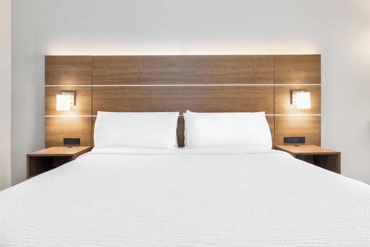 A neatly made bed with a wooden headboard, two bedside tables with wall-mounted lamps, and a plain white wall.