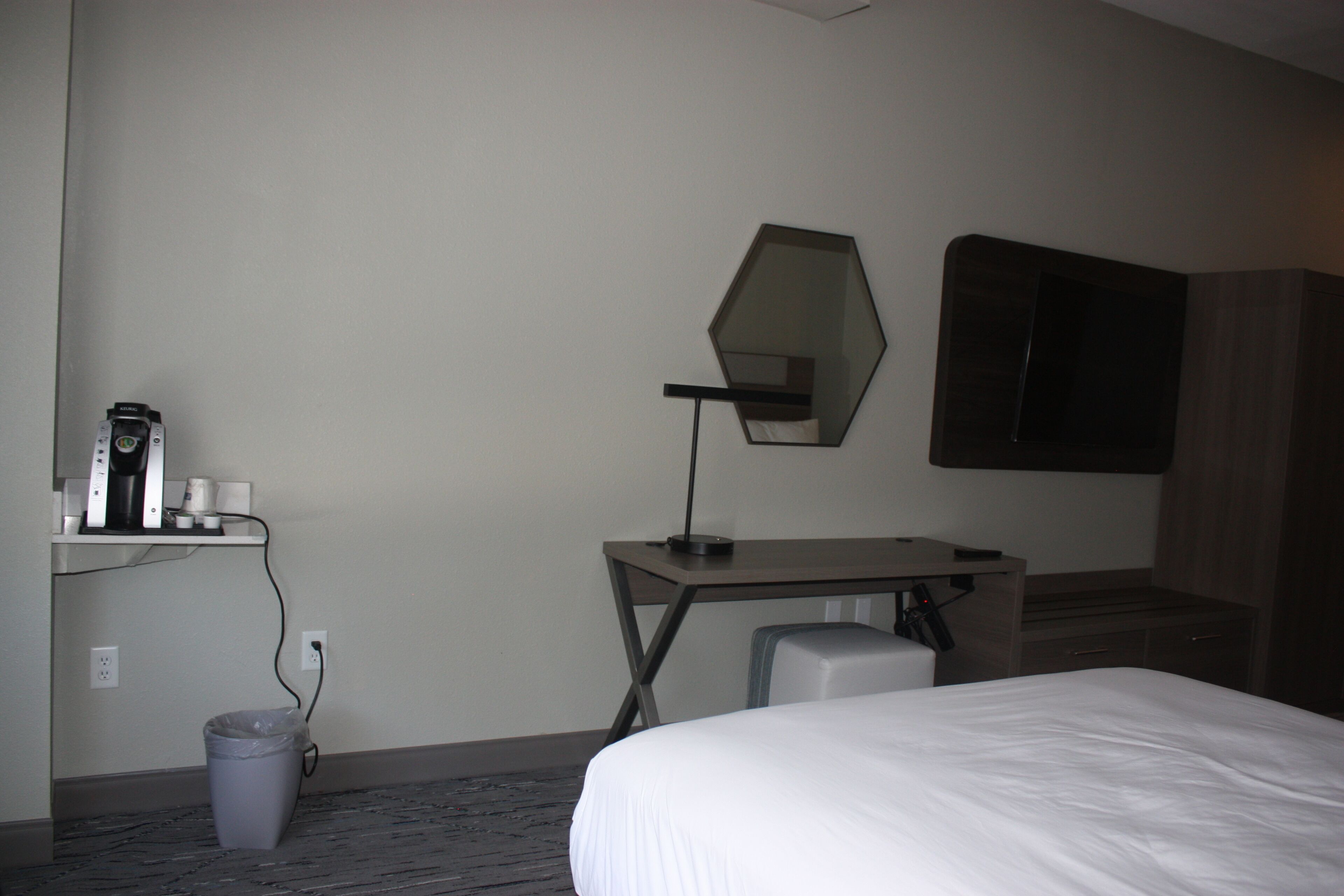 Una habitación de hotel con cama, escritorio con lámpara, un cesto de basura, espejo y armario.