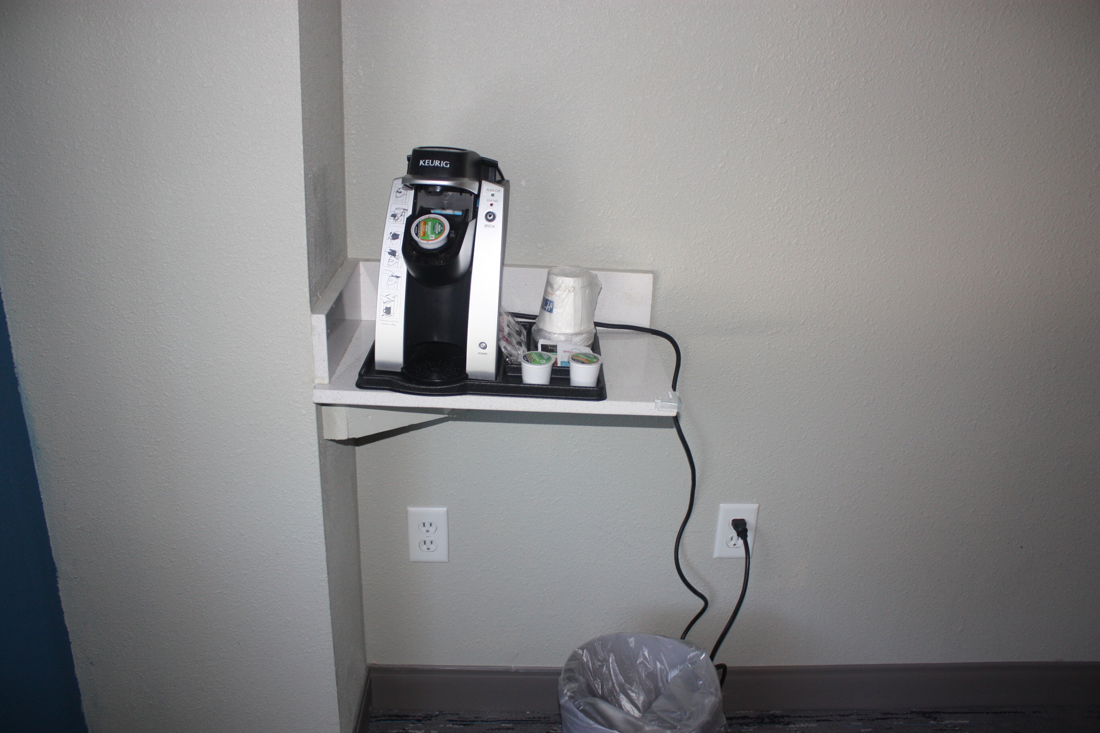 A coffee maker on a white shelf with a trash can below it.