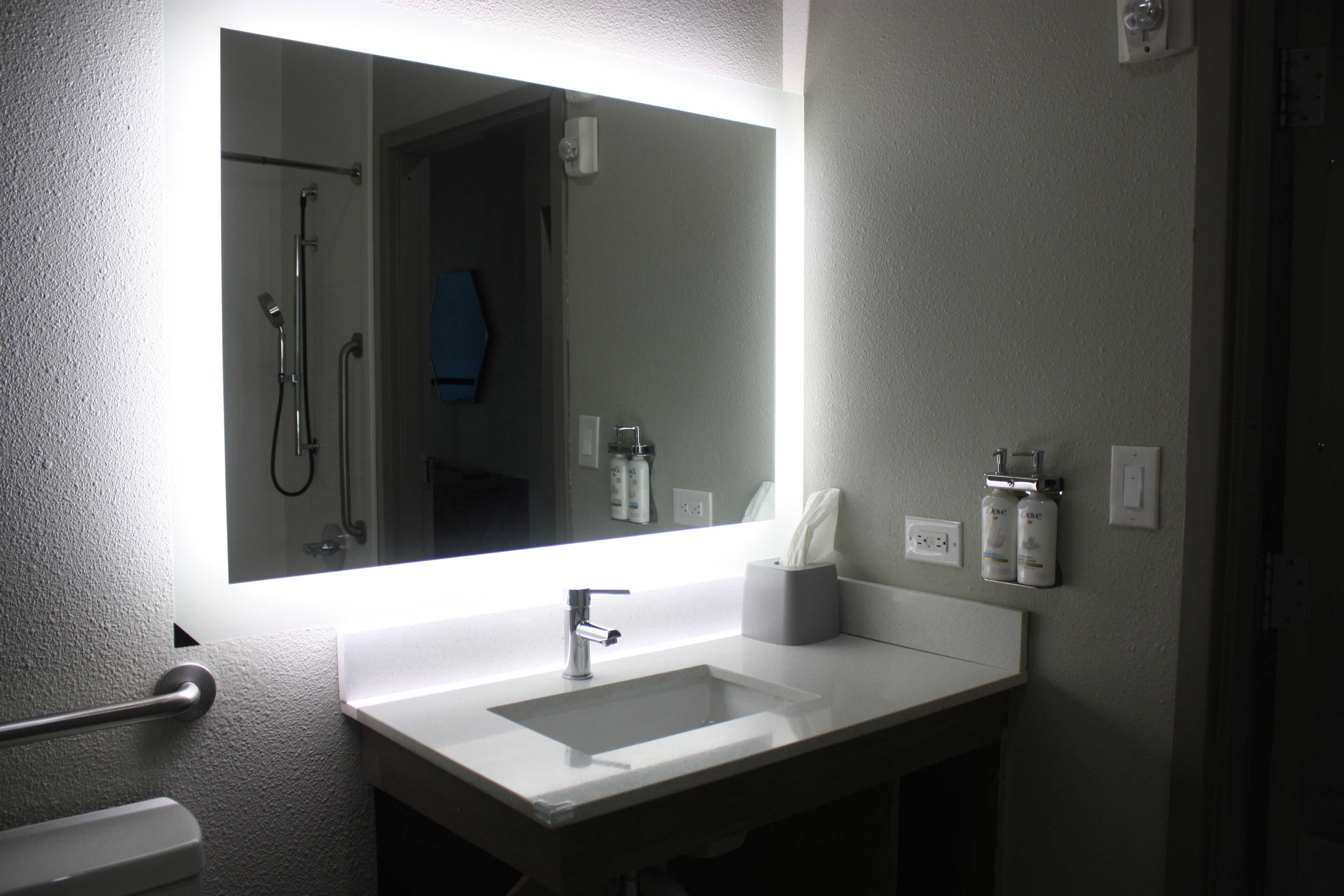 A bathroom with a large mirror, a white sink, a towel dispenser, and a shower area.
