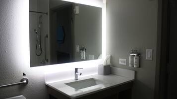 A bathroom with a large mirror, a white sink, a towel dispenser, and a shower area.
