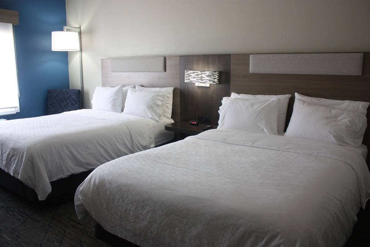 A hotel room with two beds, a nightstand, a lamp, and a wall-mounted light fixture.