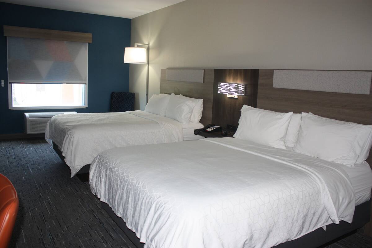 A hotel room with two beds, a nightstand, a lamp, and a window with blinds.