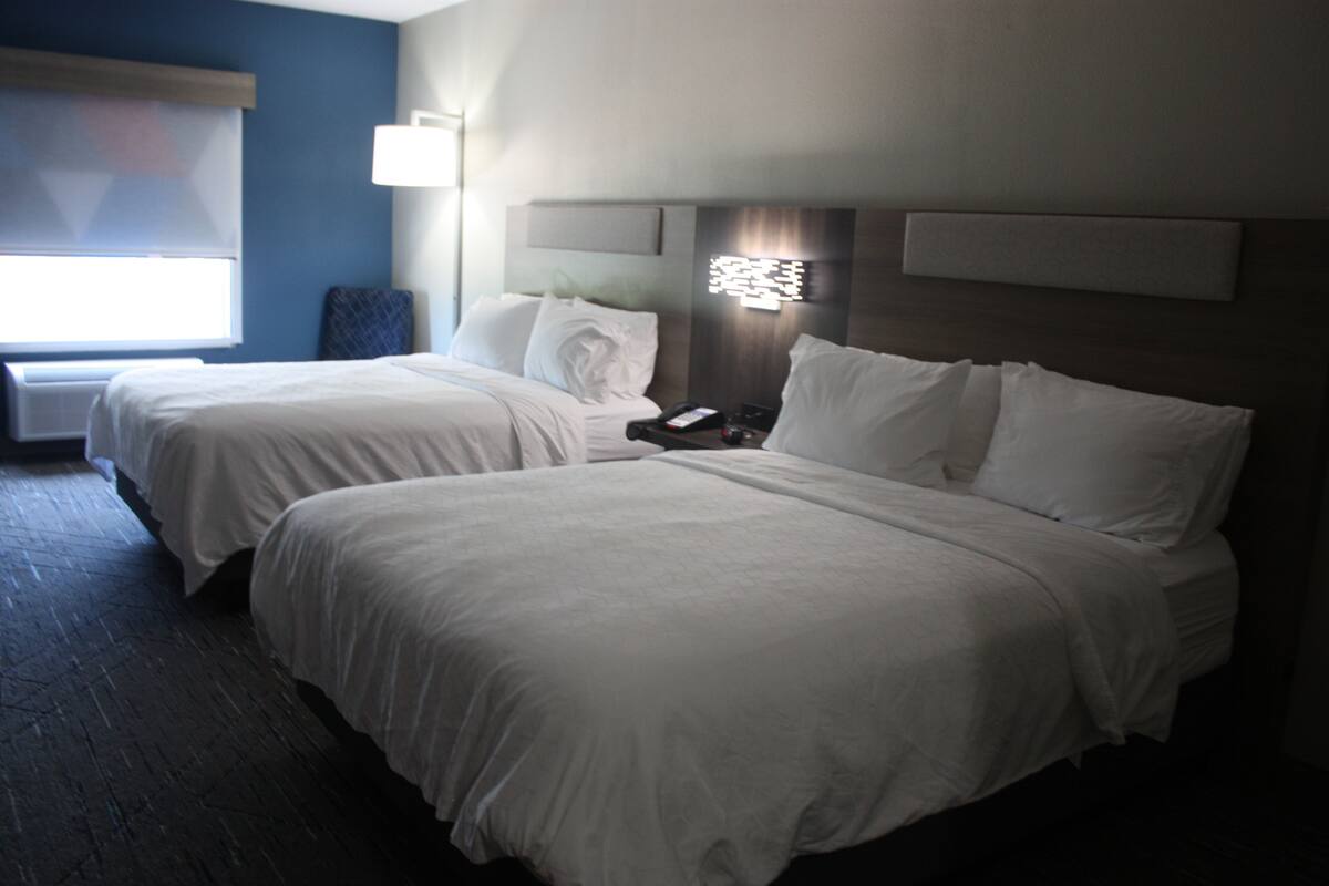 A hotel room with two beds, a bedside table, a lamp, and a window with blinds.