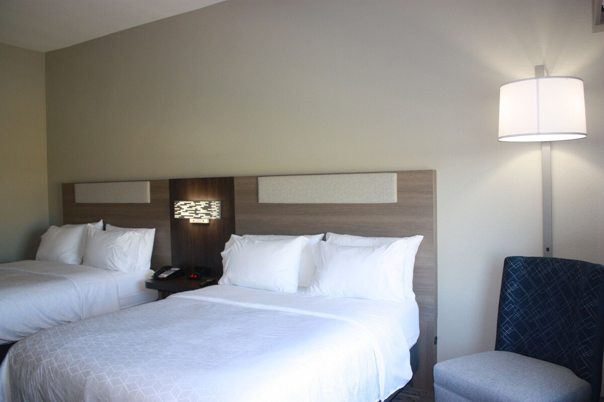 A hotel room with two beds, a nightstand with a phone, a lamp, and a chair.